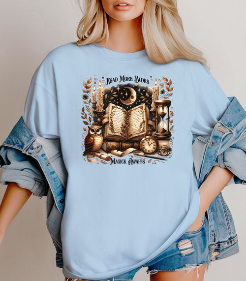 Person wearing a Chambray light blue t-shirt featuring a whimsical "Read More Books, Magick Awaits" design. The graphic showcases an open enchanted book surrounded by candles, a crescent moon, owl, pocket watch, hourglass, and magical elements, creating a mystical and literary aesthetic perfect for book lovers and fantasy fans.