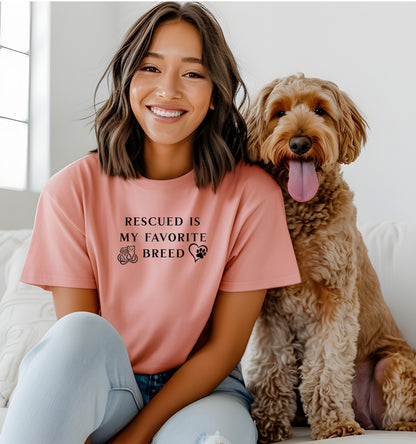 Rescued Is My Favorite Breed T-shirt featuring bold black text with small graphics of a cat, dog, and paw print heart. Printed on a sunset unisex tee, this design is perfect for animal lovers and rescue advocates. Ideal for showcasing pride in adopting pets and supporting animal rescue efforts, whether worn casually or at adoption events.