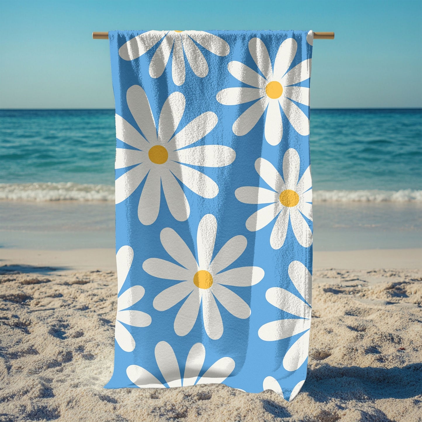 A stand holding up a vibrant blue beach towel featuring a retro daisy pattern with large white petals and yellow centers. Set against a sunny beach and ocean backdrop, this playful floral design brings cheerful vintage vibes, perfect for summer days, beach trips, and poolside lounging.