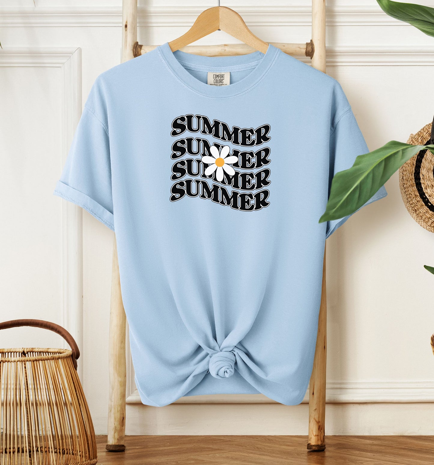 Chambray light blue T-shirt featuring a retro wavy text design that repeats the word “SUMMER” four times in bold black vintage-style letters. A large white daisy with a yellow center overlaps the middle of the design, adding a cheerful floral touch. Perfect for sunny days, vacations, and casual outings, this shirt has a nostalgic, laid-back summer vibe.