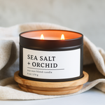 Sea Salt + Orchid soy wax blend candle in a 8 oz black matte candle tin on a wooden coaster with a neutral background