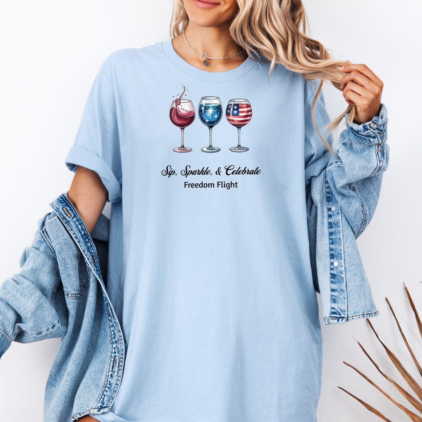 Baby blue t-shirt featuring a festive 'Sip, Sparkle, & Celebrate – Freedom Flight' design with three wine glasses: one red with sparkles, one blue with stars, and one decorated with the American flag. Perfect for 4th of July, patriotic events, summer parties, and wine lovers celebrating in style.