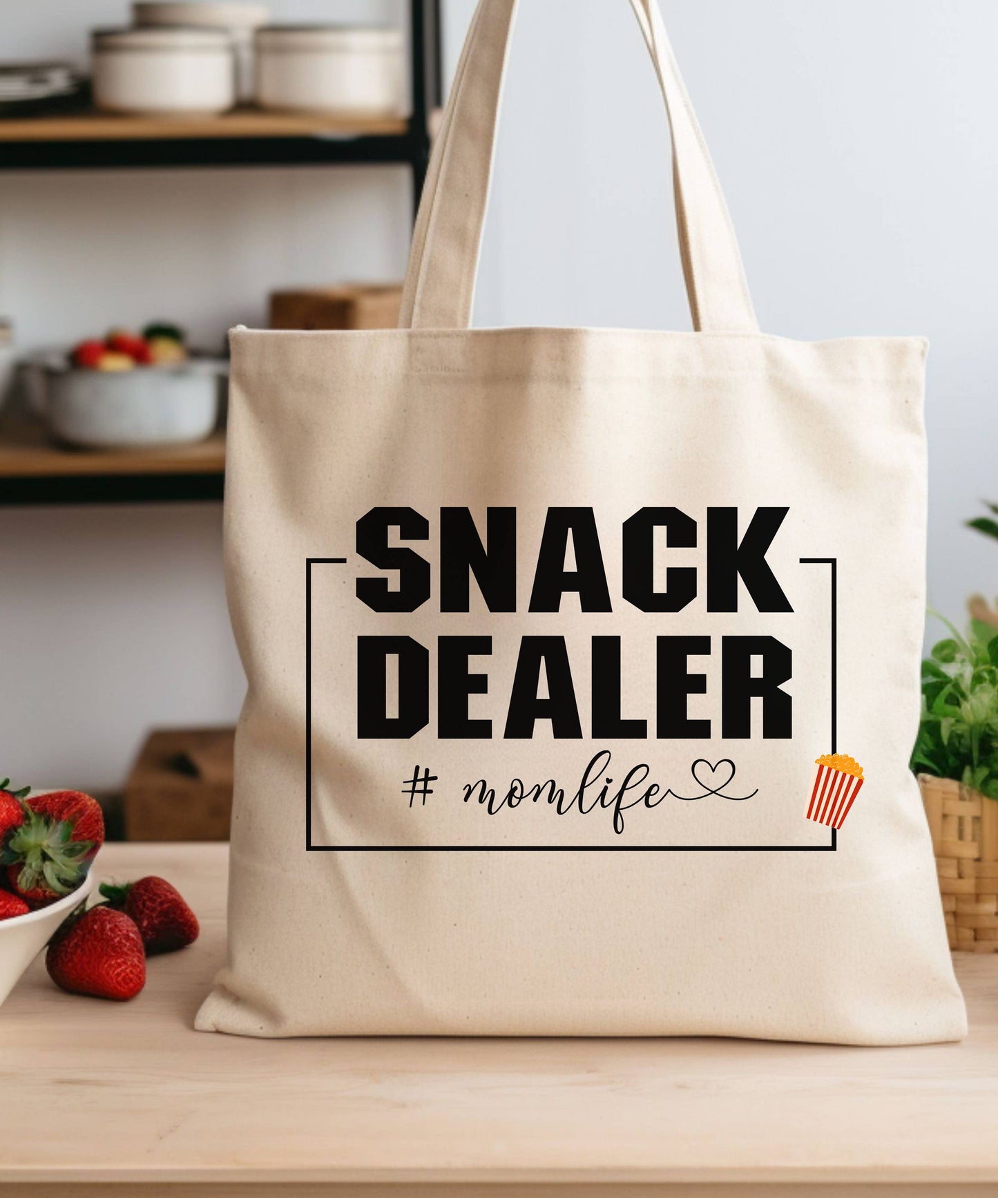 Beige tote bag with 'Snack Dealer #momlife' text on a kitchen counter with strawberries.