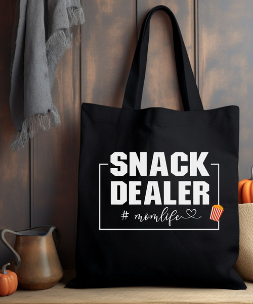 A Black tote bag with 'SNACK DEALER #momlife' text sitting on a wooden surface with pumpkins.
