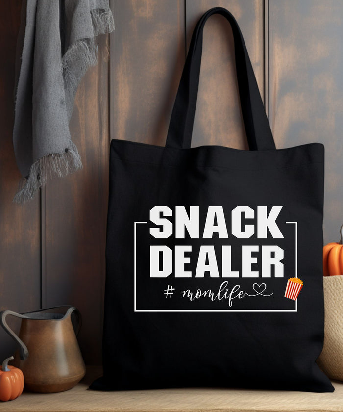 A Black tote bag with 'SNACK DEALER #momlife' text sitting on a wooden surface with pumpkins.