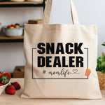 A natural tote bag with 'Snack Dealer #momlife' text sitting on a kitchen counter with strawberries next to it.