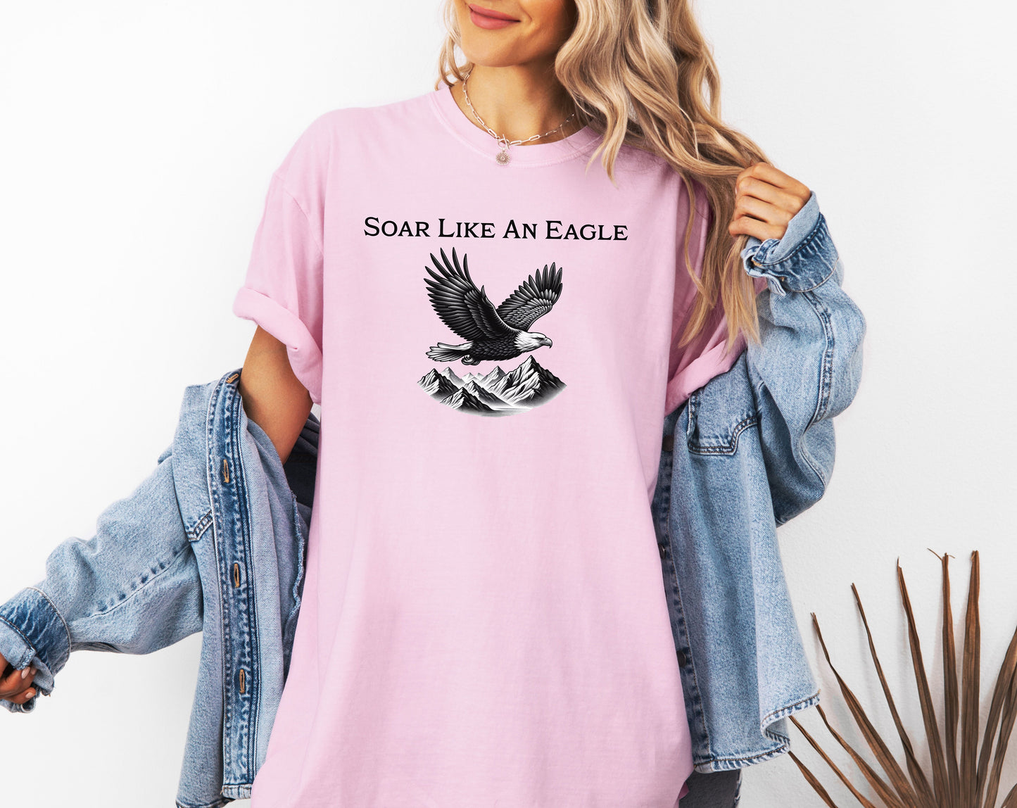 Blossom pink T-shirt featuring a bold “Soar Like An Eagle” design with white text above a detailed black and white illustration of a soaring eagle over rugged mountain peaks. The graphic captures strength, freedom, and nature’s beauty, making it perfect for outdoor enthusiasts, adventure lovers, and patriotic spirits. Modeled in a casual, relaxed fit for everyday wear.
