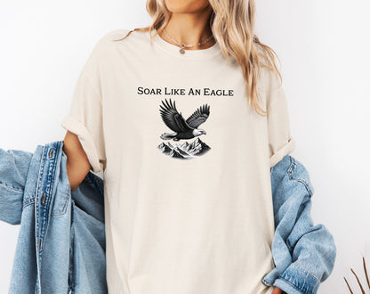 Ivory T-shirt featuring a bold “Soar Like An Eagle” design with white text above a detailed black and white illustration of a soaring eagle over rugged mountain peaks. The graphic captures strength, freedom, and nature’s beauty, making it perfect for outdoor enthusiasts, adventure lovers, and patriotic spirits. Modeled in a casual, relaxed fit for everyday wear.