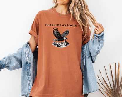 Yam T-shirt featuring a bold “Soar Like An Eagle” design with white text above a detailed black and white illustration of a soaring eagle over rugged mountain peaks. The graphic captures strength, freedom, and nature’s beauty, making it perfect for outdoor enthusiasts, adventure lovers, and patriotic spirits. Modeled in a casual, relaxed fit for everyday wear.
