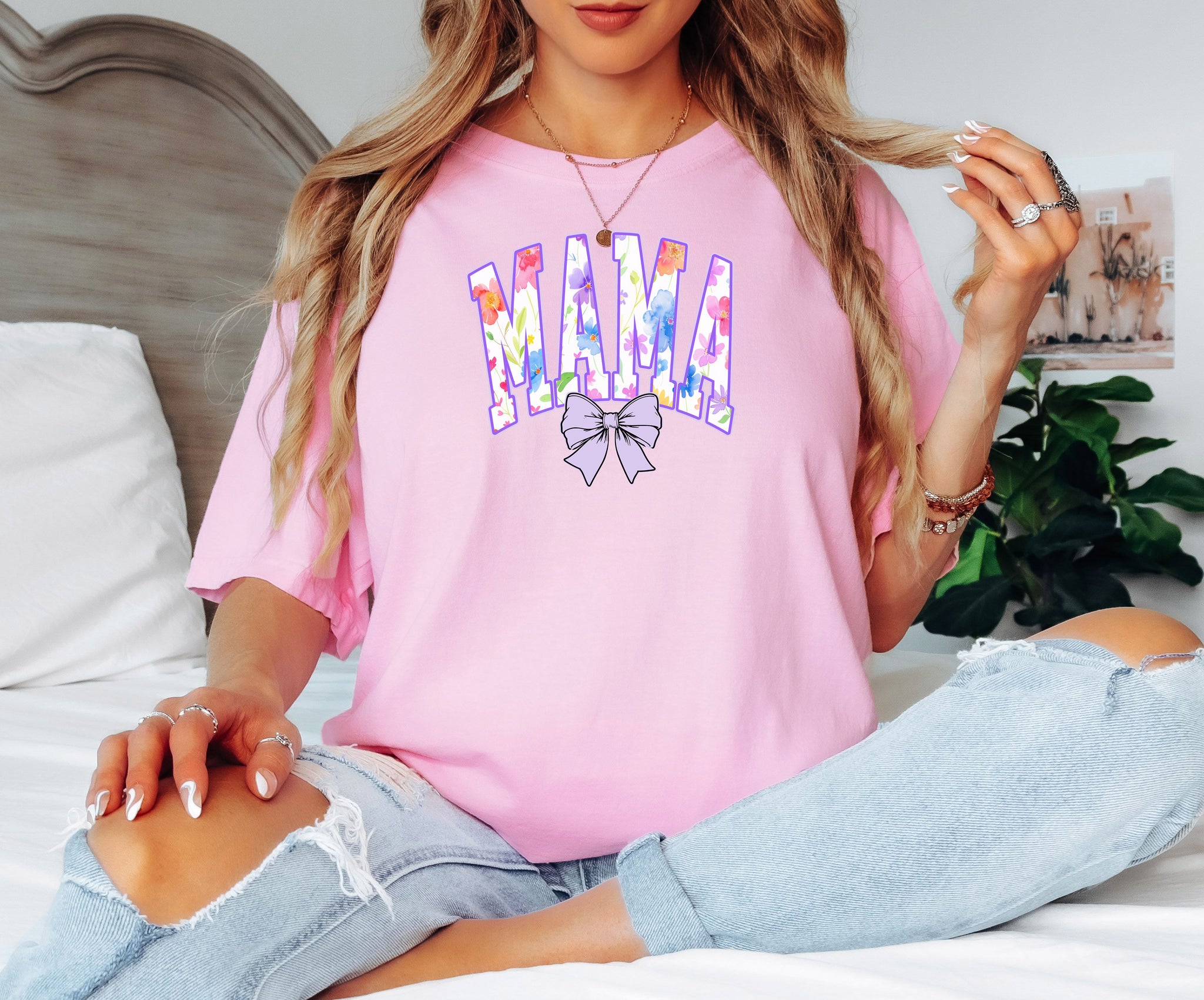 Person wearing a blossom pink T-shirt featuring a bold “MAMA” design in large block letters filled with a vibrant violet, pink, orange, yellow, and blue floral pattern outlined in purple. A soft violet coquette bow sits below the text, adding a feminine touch. This spring-inspired graphic tee blends floral elegance with playful charm, perfect for moms who love colorful and trendy fashion.