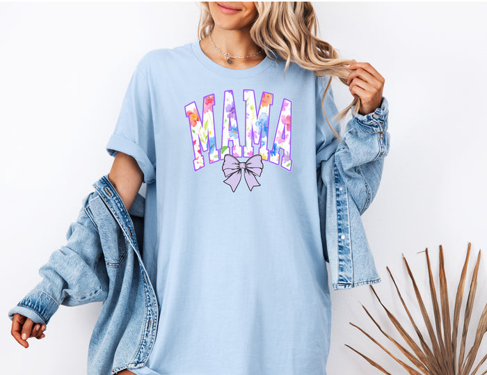 Person wearing a chambray light blue T-shirt featuring a bold “MAMA” design in large block letters filled with a vibrant violet, pink, orange, yellow, and blue floral pattern outlined in purple. A soft violet coquette bow sits below the text, adding a feminine touch.  This spring-inspired graphic tee blends floral elegance with playful charm, perfect for moms who love colorful and trendy fashion.