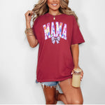 Person wearing a chili T-shirt featuring a bold “MAMA” design in large block letters filled with a vibrant violet, pink, orange, yellow, and blue floral pattern outlined in purple. A soft violet coquette bow sits below the text, adding a feminine touch. 
This spring-inspired graphic tee blends floral elegance with playful charm, perfect for moms who love colorful and trendy fashion.