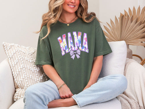 Person wearing a hemp color T-shirt featuring a bold “MAMA” design in large block letters filled with a vibrant violet, pink, orange, yellow, and blue floral pattern outlined in purple. A soft violet coquette bow sits below the text, adding a feminine touch.  This spring-inspired graphic tee blends floral elegance with playful charm, perfect for moms who love colorful and trendy fashion.