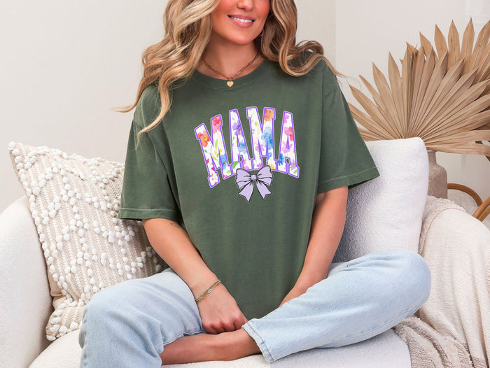 Person wearing a hemp color T-shirt featuring a bold “MAMA” design in large block letters filled with a vibrant violet, pink, orange, yellow, and blue floral pattern outlined in purple. A soft violet coquette bow sits below the text, adding a feminine touch.  This spring-inspired graphic tee blends floral elegance with playful charm, perfect for moms who love colorful and trendy fashion.