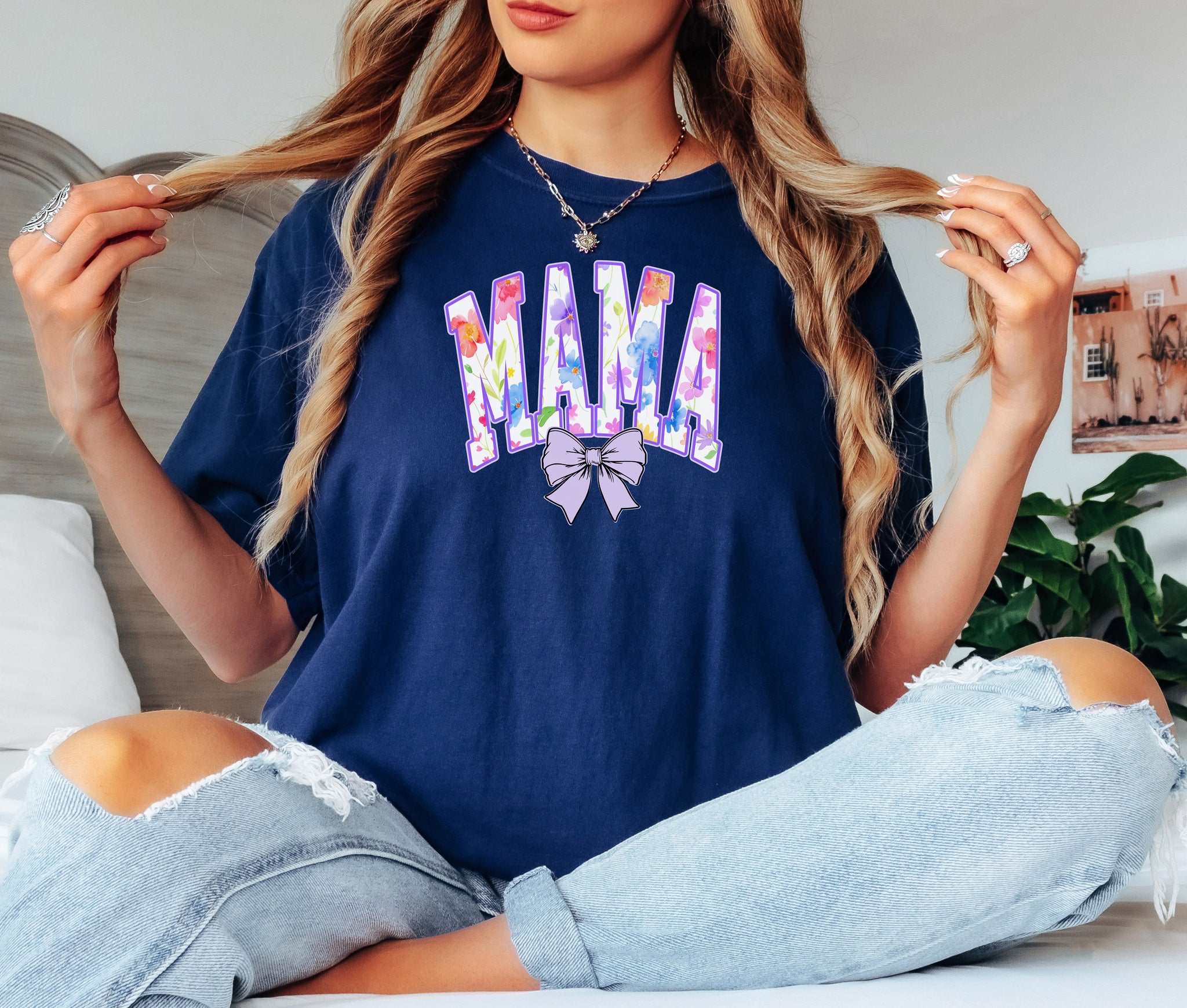 Person wearing a true navy T-shirt featuring a bold “MAMA” design in large block letters filled with a vibrant violet, pink, orange, yellow, and blue floral pattern outlined in purple. A soft violet coquette bow sits below the text, adding a feminine touch.  This spring-inspired graphic tee blends floral elegance with playful charm, perfect for moms who love colorful and trendy fashion.