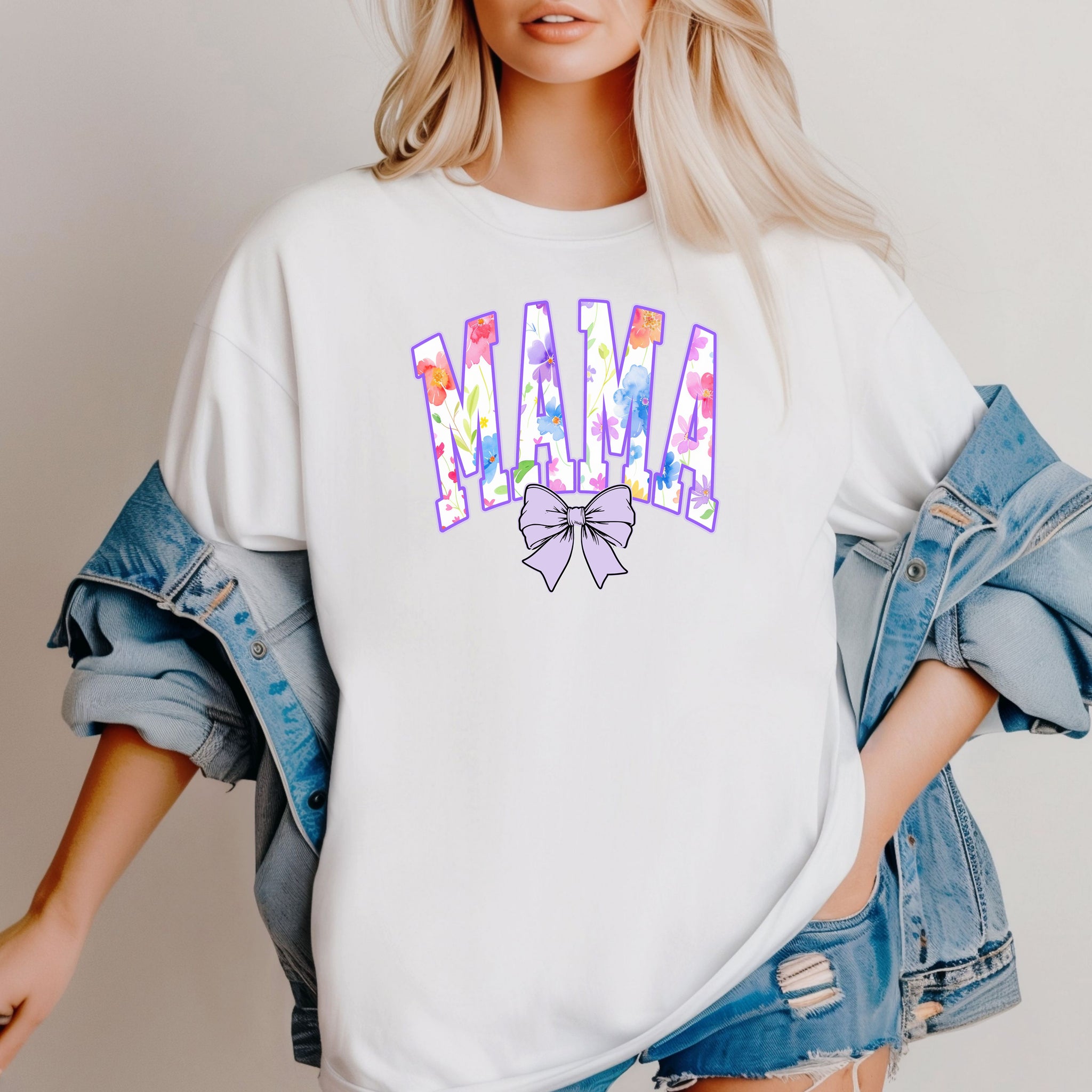 Person wearing a classic white T-shirt featuring a bold “MAMA” design in large block letters filled with a vibrant violet, pink, orange, yellow, and blue floral pattern outlined in purple. A soft violet coquette bow sits below the text, adding a feminine touch.  This spring-inspired graphic tee blends floral elegance with playful charm, perfect for moms who love colorful and trendy fashion.