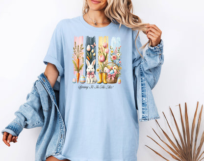 Spring Is In The Air T-shirt in soft baby blue featuring a charming Sprint and Easter design with a white bunny wearing a pink coquette bow, colorful tulips, carrots, pastel eggs, rain boots, and a floral basket. The background includes vertical pastel paint strokes in pink, yellow, blue, and navy, adding a cheerful seasonal touch perfect for spring and Easter celebrations.