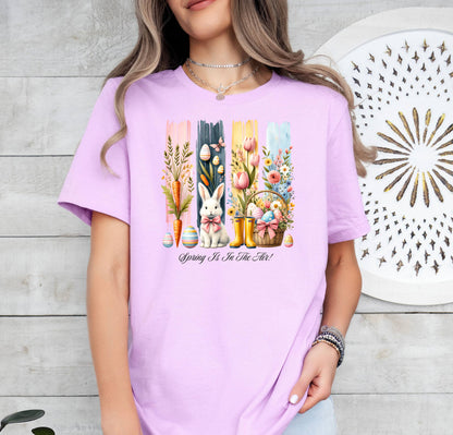Spring Is In The Air T-shirt in soft lilac featuring a charming Sprint and Easter design with a white bunny wearing a pink coquette bow, colorful tulips, carrots, pastel eggs, rain boots, and a floral basket. The background includes vertical pastel paint strokes in pink, yellow, blue, and navy, adding a cheerful seasonal touch perfect for spring and Easter celebrations.