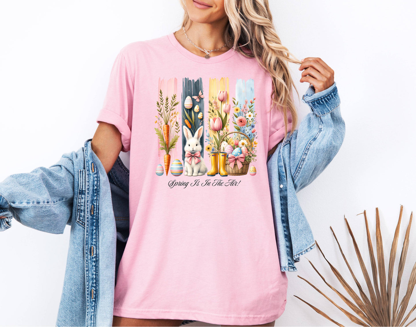 Spring Is In The Air T-shirt in pink featuring a charming Sprint and Easter design with a white bunny wearing a pink coquette bow, colorful tulips, carrots, pastel eggs, rain boots, and a floral basket. The background includes vertical pastel paint strokes in pink, yellow, blue, and navy, adding a cheerful seasonal touch perfect for spring and Easter celebrations.