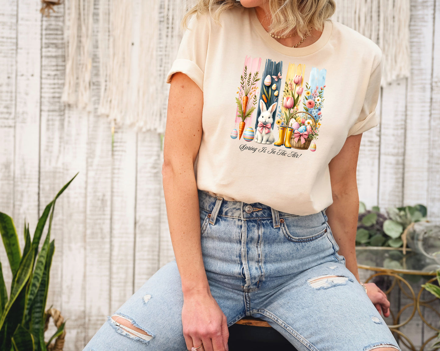 Spring Is In The Air T-shirt in soft cream featuring a charming Sprint and Easter design with a white bunny wearing a pink coquette bow, colorful tulips, carrots, pastel eggs, rain boots, and a floral basket. The background includes vertical pastel paint strokes in pink, yellow, blue, and navy, adding a cheerful seasonal touch perfect for spring and Easter celebrations.