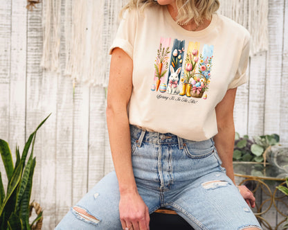 Spring Is In The Air T-shirt in soft cream featuring a charming Sprint and Easter design with a white bunny wearing a pink coquette bow, colorful tulips, carrots, pastel eggs, rain boots, and a floral basket. The background includes vertical pastel paint strokes in pink, yellow, blue, and navy, adding a cheerful seasonal touch perfect for spring and Easter celebrations.