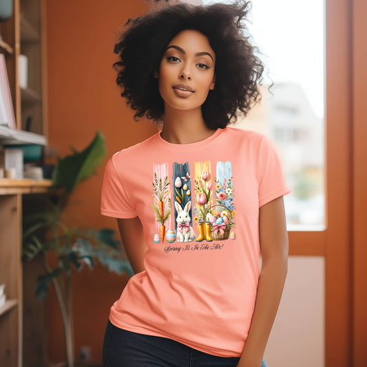 Spring Is In The Air T-shirt in sunset featuring a charming Sprint and Easter design with a white bunny wearing a pink coquette bow, colorful tulips, carrots, pastel eggs, rain boots, and a floral basket. The background includes vertical pastel paint strokes in pink, yellow, blue, and navy, adding a cheerful seasonal touch perfect for spring and Easter celebrations.