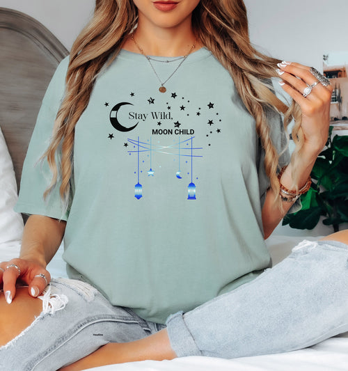 Stay Wild Moon Child T-shirt featuring a celestial design with a crescent moon, stars, and hanging lanterns. The text 'Stay Wild, Moon Child' is surrounded by cosmic elements, evoking a dreamy bohemian vibe. Printed on a bay unisex tee, this mystical and free-spirited graphic is perfect for astrology lovers, night sky dreamers, and those who embrace their wild, cosmic side.