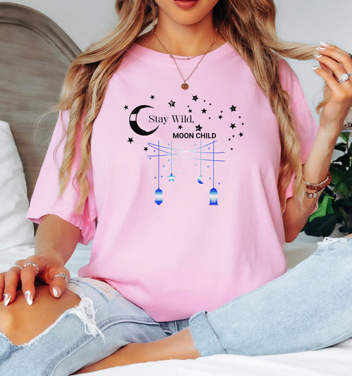 Stay Wild Moon Child T-shirt featuring a celestial design with a crescent moon, stars, and hanging lanterns. The text 'Stay Wild, Moon Child' is surrounded by cosmic elements, evoking a dreamy bohemian vibe. Printed on a blossom pink unisex tee, this mystical and free-spirited graphic is perfect for astrology lovers, night sky dreamers, and those who embrace their wild, cosmic side.