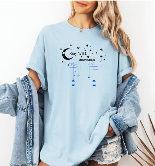 Stay Wild Moon Child T-shirt featuring a celestial design with a crescent moon, stars, and hanging lanterns. The text 'Stay Wild, Moon Child' is surrounded by cosmic elements, evoking a dreamy bohemian vibe. Printed on a chambray light blue unisex tee, this mystical and free-spirited graphic is perfect for astrology lovers, night sky dreamers, and those who embrace their wild, cosmic side.