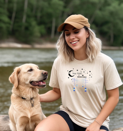 Stay Wild Moon Child T-shirt featuring a celestial design with a crescent moon, stars, and hanging lanterns. The text 'Stay Wild, Moon Child' is surrounded by cosmic elements, evoking a dreamy bohemian vibe. Printed on an Ivory unisex tee, this mystical and free-spirited graphic is perfect for astrology lovers, night sky dreamers, and those who embrace their wild, cosmic side.