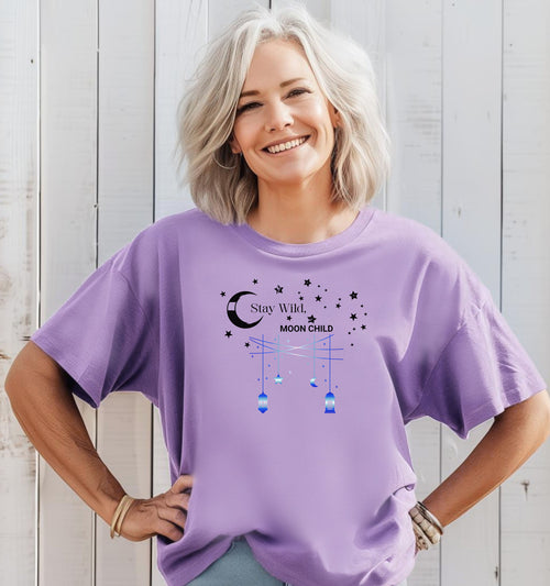 Stay Wild Moon Child T-shirt featuring a celestial design with a crescent moon, stars, and hanging lanterns. The text 'Stay Wild, Moon Child' is surrounded by cosmic elements, evoking a dreamy bohemian vibe. Printed on a violet unisex tee, this mystical and free-spirited graphic is perfect for astrology lovers, night sky dreamers, and those who embrace their wild, cosmic side.