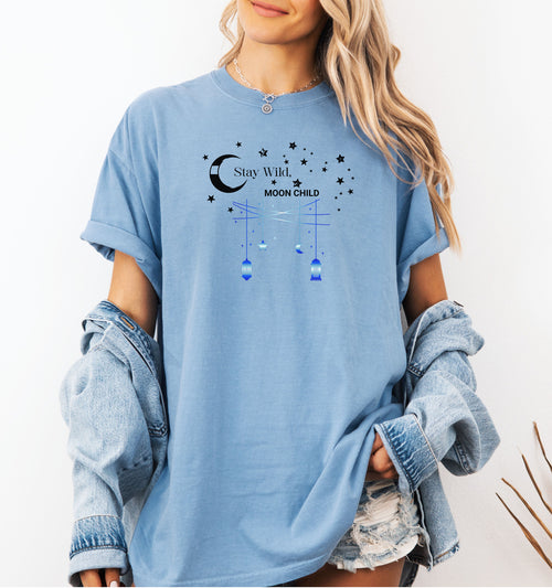 Stay Wild Moon Child T-shirt featuring a celestial design with a crescent moon, stars, and hanging lanterns. The text 'Stay Wild, Moon Child' is surrounded by cosmic elements, evoking a dreamy bohemian vibe. Printed on a washed denim unisex tee, this mystical and free-spirited graphic is perfect for astrology lovers, night sky dreamers, and those who embrace their wild, cosmic side.