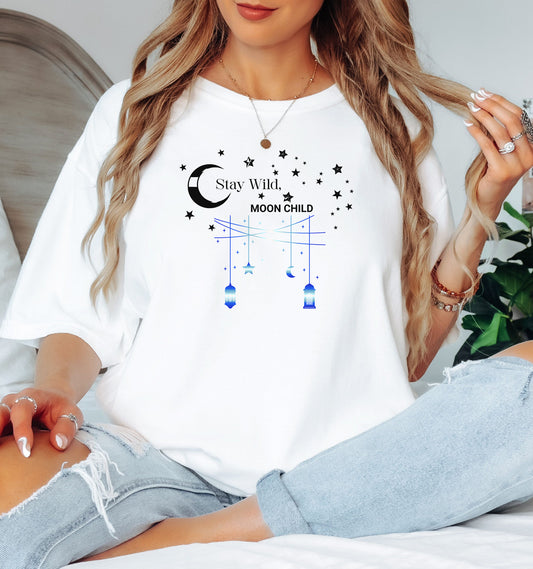 Stay Wild Moon Child T-shirt featuring a celestial design with a crescent moon, stars, and hanging lanterns. The text 'Stay Wild, Moon Child' is surrounded by cosmic elements, evoking a dreamy bohemian vibe. Printed on a white unisex tee, this mystical and free-spirited graphic is perfect for astrology lovers, night sky dreamers, and those who embrace their wild, cosmic side.