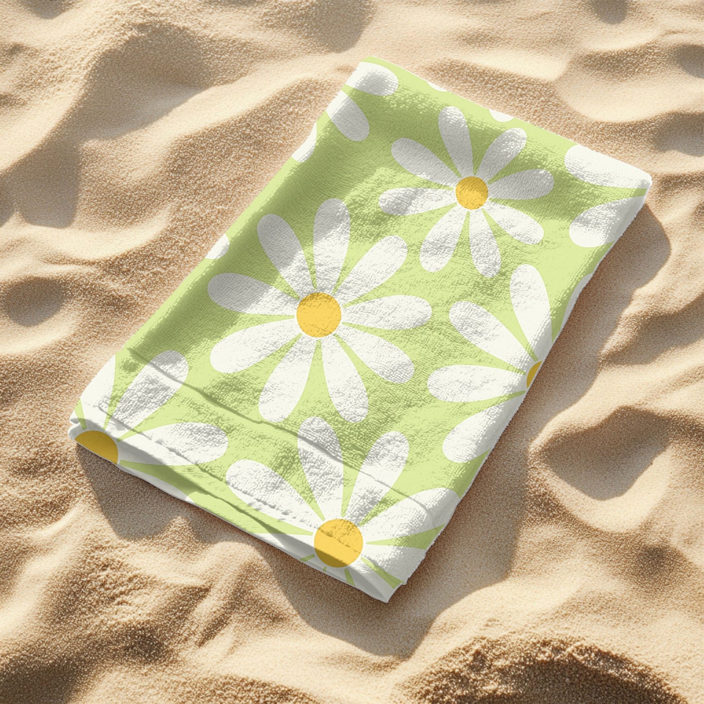 Light green folded beach towel laying on the sand featuring a cheerful retro daisy pattern with large white petals and yellow centers. Set on a sunny beach with ocean waves in the background and sunglasses in the sand, this towel brings playful vintage vibes to any seaside or poolside day.