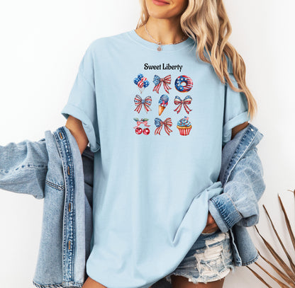 Chambray light blue t-shirt featuring a festive 'Sweet Liberty' design with patriotic icons including bows, cherries, a donut, ice cream cone, and cupcake decorated in red, white, and blue stars and stripes. Fun, colorful 4th of July and summer celebration shirt perfect for parties, barbecues, and casual wear.