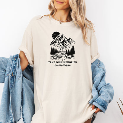 Ivory unisex T-shirt featuring a black minimalist outdoor design of mountains, pine trees, and a winding river under a crescent moon. Below the scene, the text reads “Take Only Memories, Leave Only Footprints” in elegant script and bold font. Modeled with a casual denim jacket for a relaxed, nature-inspired style.