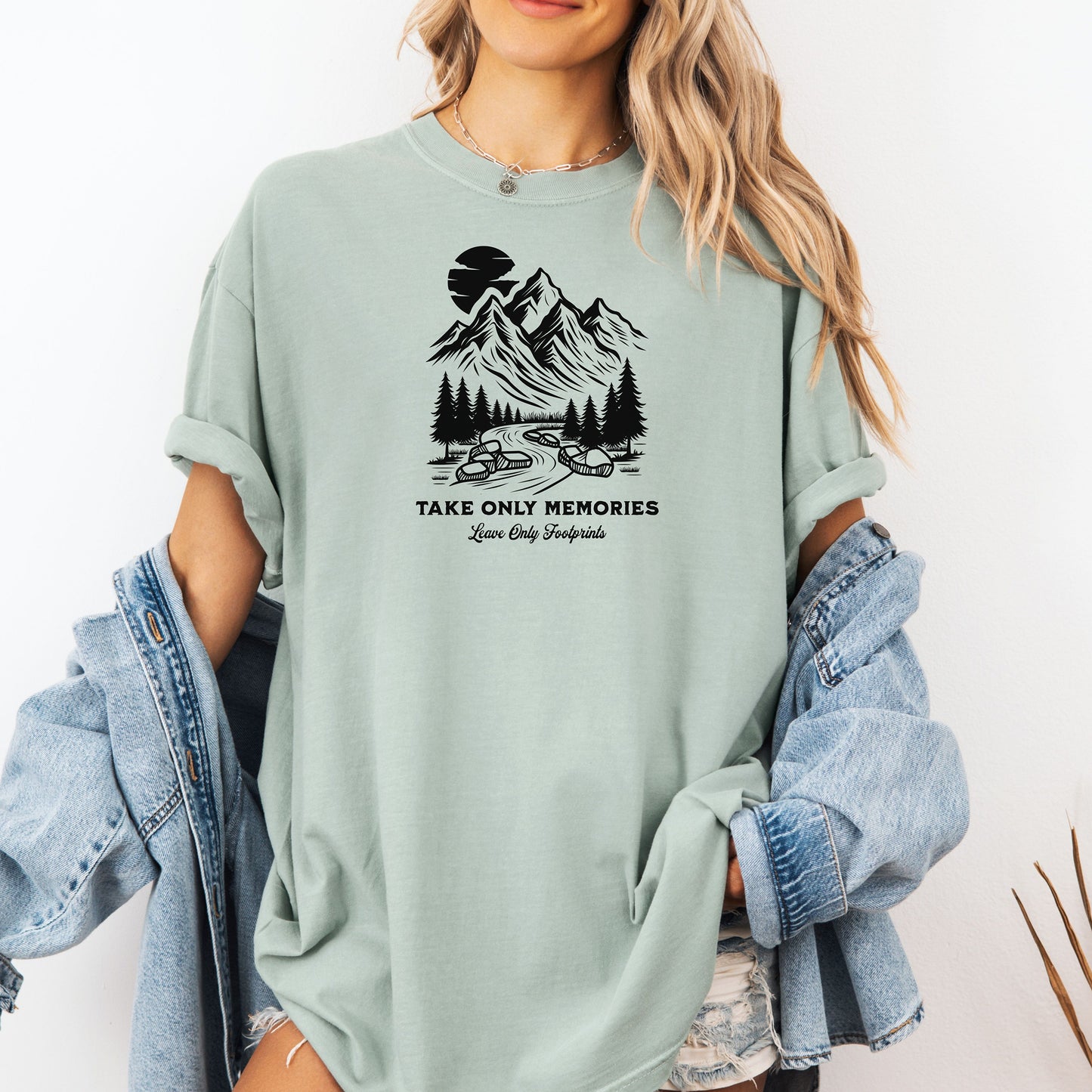 Bay light green unisex T-shirt featuring a black minimalist outdoor design of mountains, pine trees, and a winding river under a crescent moon. Below the scene, the text reads “Take Only Memories, Leave Only Footprints” in elegant script and bold font. 