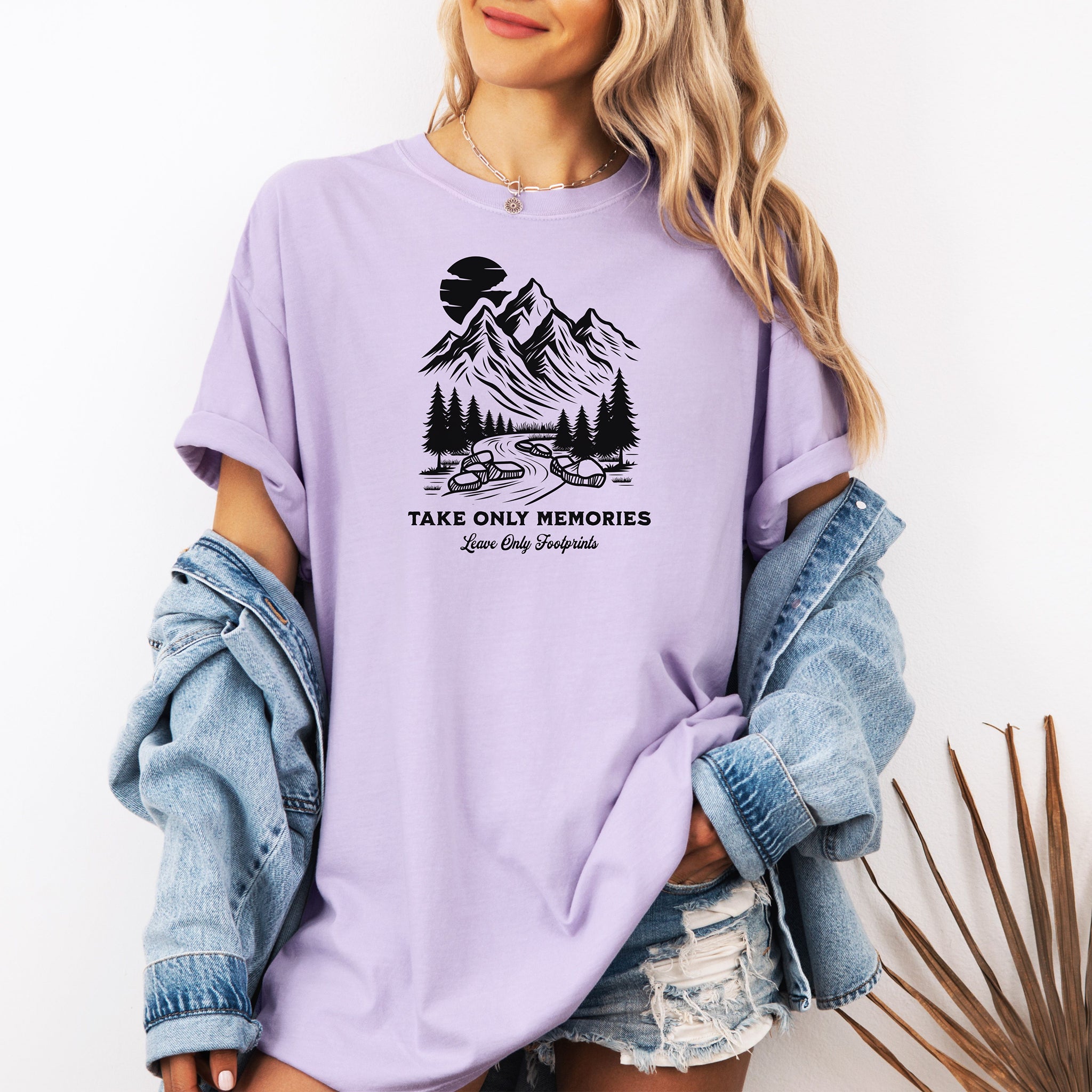 Orchid unisex T-shirt featuring a black minimalist outdoor design of mountains, pine trees, and a winding river under a crescent moon. Below the scene, the text reads “Take Only Memories, Leave Only Footprints” in elegant script and bold font. Modeled with a casual denim jacket for a relaxed, nature-inspired style.