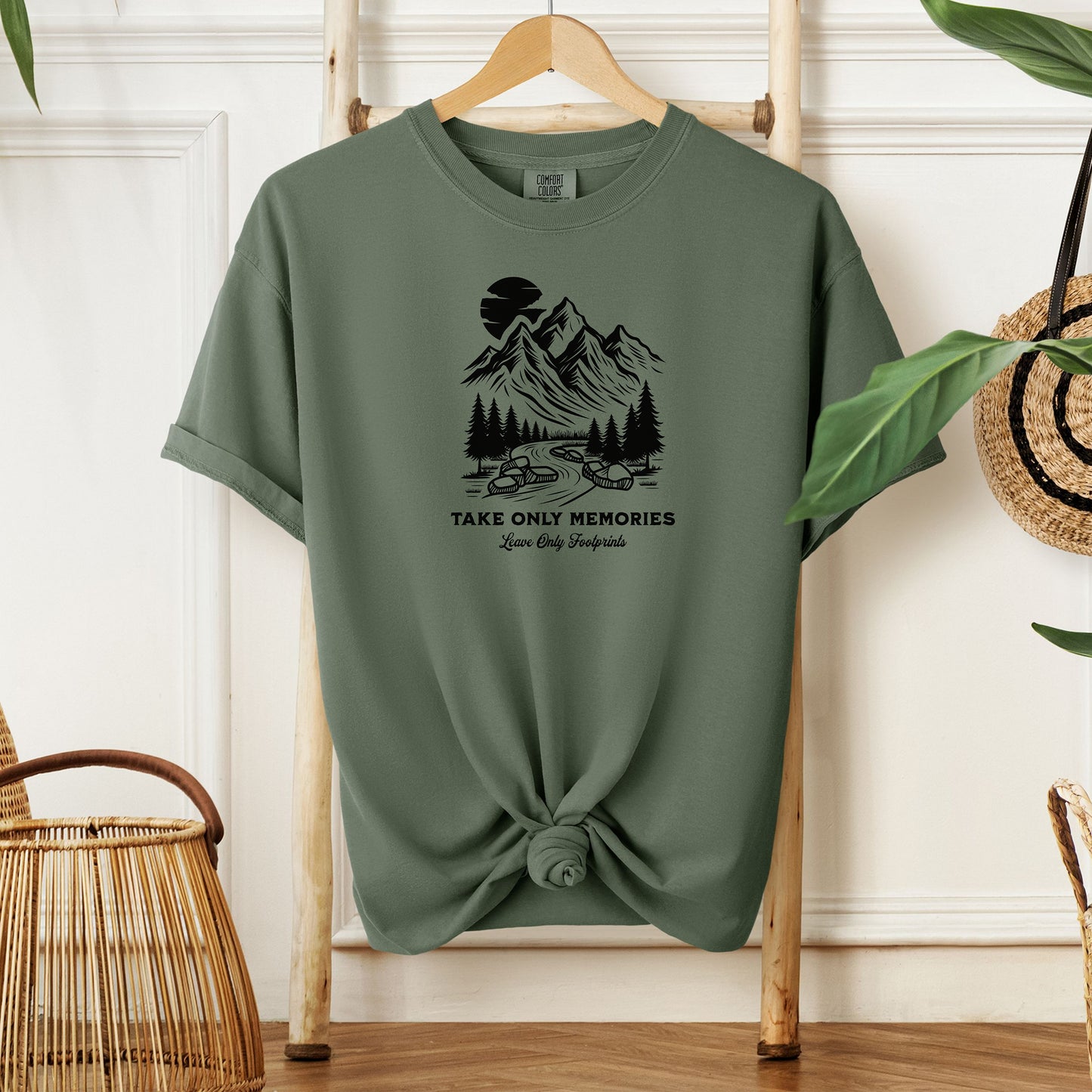 Sage unisex T-shirt featuring a black minimalist outdoor design of mountains, pine trees, and a winding river under a crescent moon. Below the scene, the text reads “Take Only Memories, Leave Only Footprints” in elegant script and bold font. Modeled with a casual denim jacket for a relaxed, nature-inspired style.