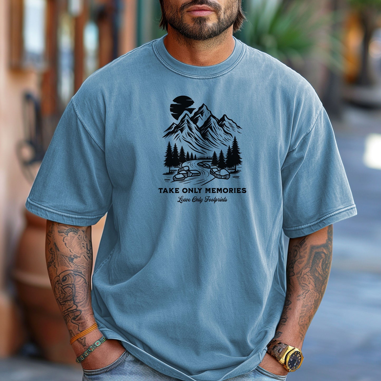 Washed denim unisex T-shirt featuring a black minimalist outdoor design of mountains, pine trees, and a winding river under a crescent moon. Below the scene, the text reads “Take Only Memories, Leave Only Footprints” in elegant script and bold font. Modeled with a casual denim jacket for a relaxed, nature-inspired style.