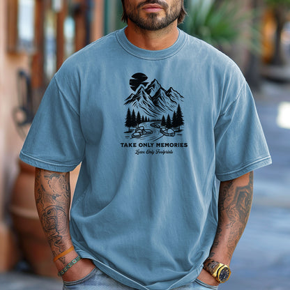 Washed denim unisex T-shirt featuring a black minimalist outdoor design of mountains, pine trees, and a winding river under a crescent moon. Below the scene, the text reads “Take Only Memories, Leave Only Footprints” in elegant script and bold font. Modeled with a casual denim jacket for a relaxed, nature-inspired style.