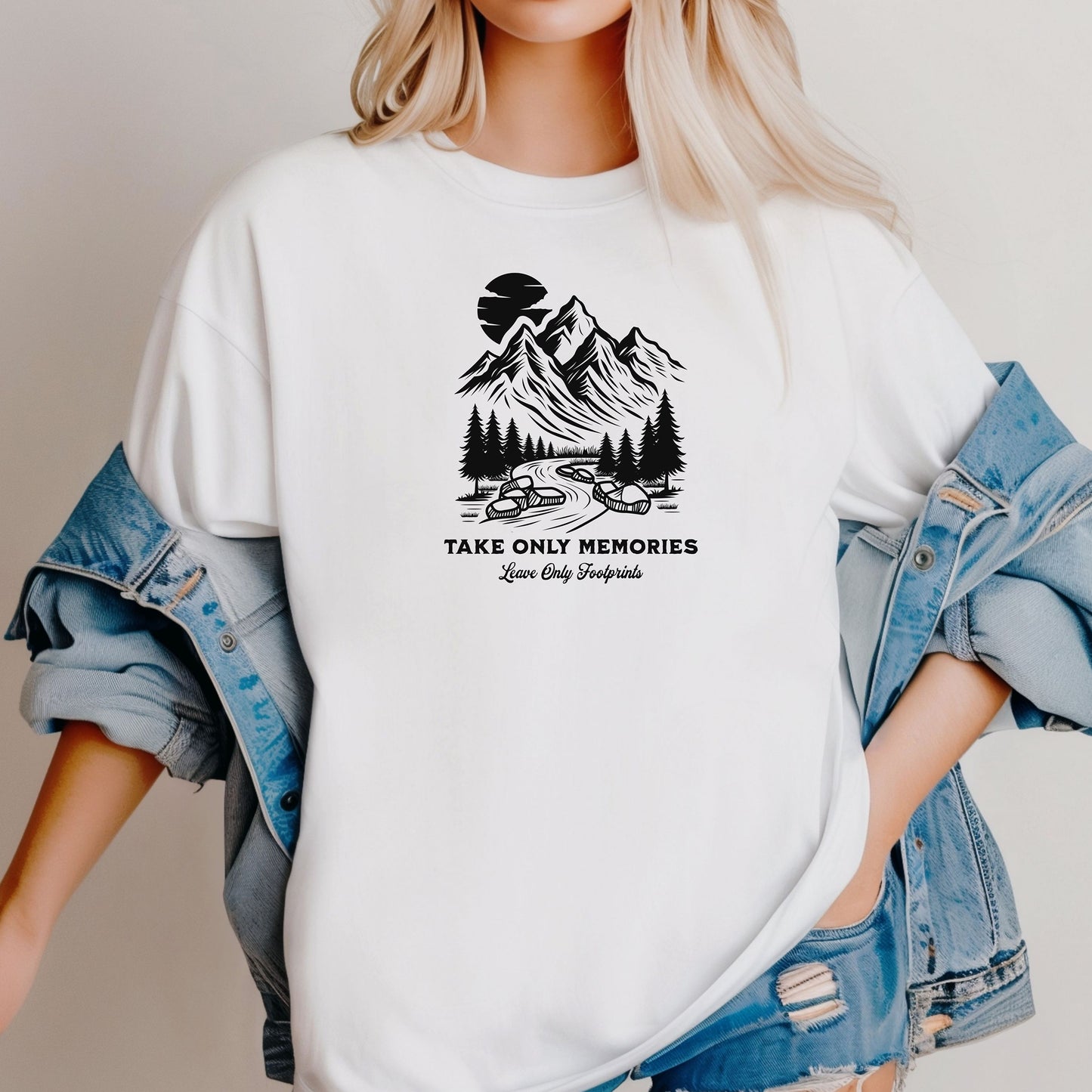 White unisex T-shirt featuring a black minimalist outdoor design of mountains, pine trees, and a winding river under a crescent moon. Below the scene, the text reads “Take Only Memories, Leave Only Footprints” in elegant script and bold font. Modeled with a casual denim jacket for a relaxed, nature-inspired style.