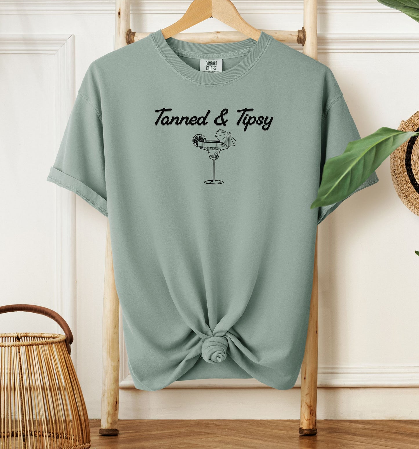 Bay green T-shirt featuring the fun phrase “Tanned & Tipsy” with a simple line art illustration of a margarita glass garnished with a lime wedge and a tiny cocktail umbrella. Perfect for summer beach parties, vacations, or casual outings, this relaxed and playful shirt embodies sunny vibes and carefree fun.