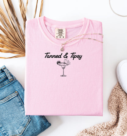 Blossom pink T-shirt featuring the fun phrase “Tanned & Tipsy” with a simple line art illustration of a margarita glass garnished with a lime wedge and a tiny cocktail umbrella. Perfect for summer beach parties, vacations, or casual outings, this relaxed and playful shirt embodies sunny vibes and carefree fun.