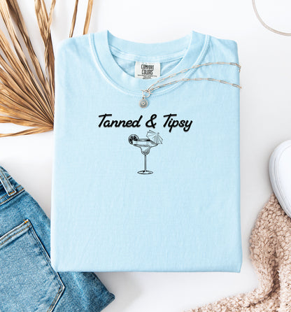 Chambray light blue T-shirt featuring the fun phrase “Tanned & Tipsy” with a simple line art illustration of a margarita glass garnished with a lime wedge and a tiny cocktail umbrella. Perfect for summer beach parties, vacations, or casual outings, this relaxed and playful shirt embodies sunny vibes and carefree fun.