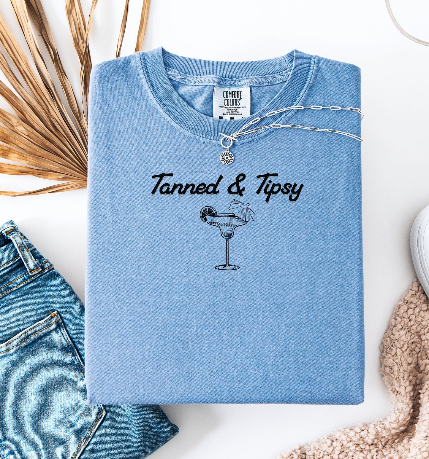 Washed Denim T-shirt featuring the fun phrase “Tanned & Tipsy” with a simple line art illustration of a margarita glass garnished with a lime wedge and a tiny cocktail umbrella. Perfect for summer beach parties, vacations, or casual outings, this relaxed and playful shirt embodies sunny vibes and carefree fun.
