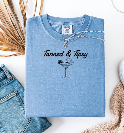 Washed Denim T-shirt featuring the fun phrase “Tanned & Tipsy” with a simple line art illustration of a margarita glass garnished with a lime wedge and a tiny cocktail umbrella. Perfect for summer beach parties, vacations, or casual outings, this relaxed and playful shirt embodies sunny vibes and carefree fun.