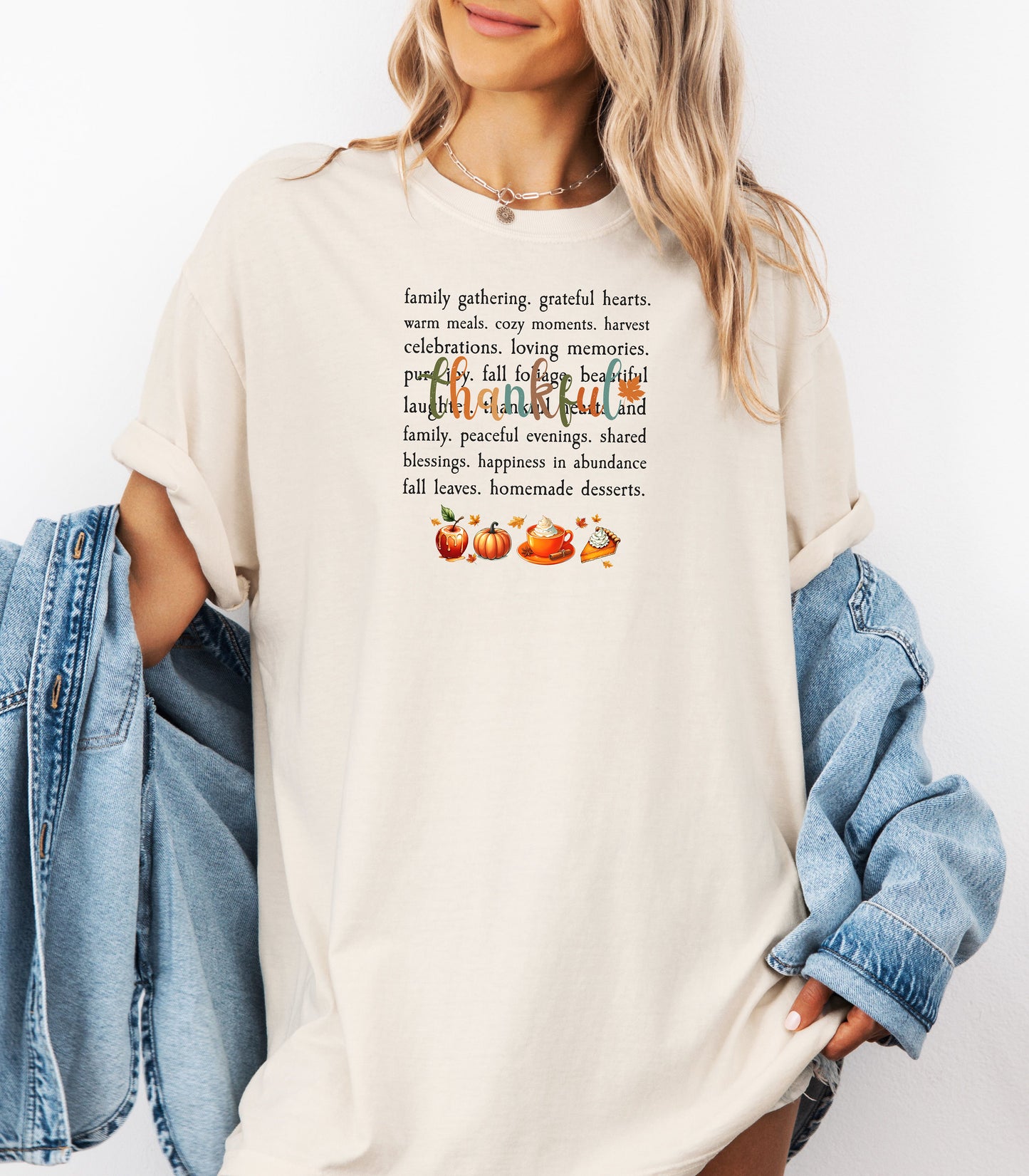 Person wearing an Ivory T-shirt with a festive “Thankful” harvest-themed design featuring fall words like family gathering, grateful hearts, cozy moments, and homemade desserts, with autumn illustrations of an apple, pumpkin, pie, and fall leaves. A warm and cheerful tee perfect for Thanksgiving, family gatherings, or celebrating the fall season.