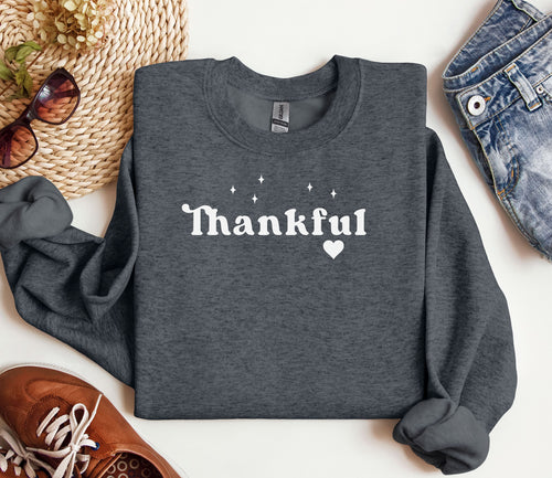 Dark Heather Gildan sweatshirt featuring the word “Thankful” in bold white retro-style font, accented with small sparkling stars above and a heart below. A simple yet meaningful design that celebrates gratitude and cozy fall vibes—perfect for Thanksgiving, autumn gatherings, or everyday wear.