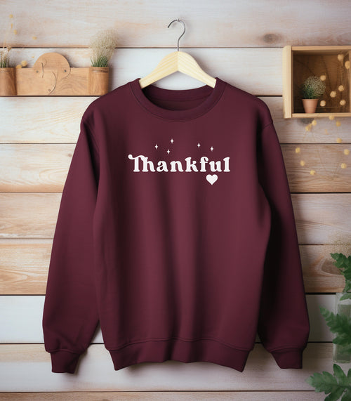 Maroon Gildan sweatshirt featuring the word “Thankful” in bold white retro-style font, accented with small sparkling stars above and a heart below. A simple yet meaningful design that celebrates gratitude and cozy fall vibes—perfect for Thanksgiving, autumn gatherings, or everyday wear.